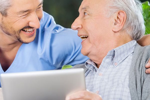 A close up of a doctor with his arm around an older man while they’re both smiling and the older man is holding an iPad