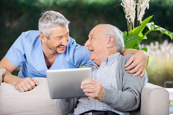 An older man sitting on a couch while holding an iPad and looking at the dentist who is smiling with his arm around the man