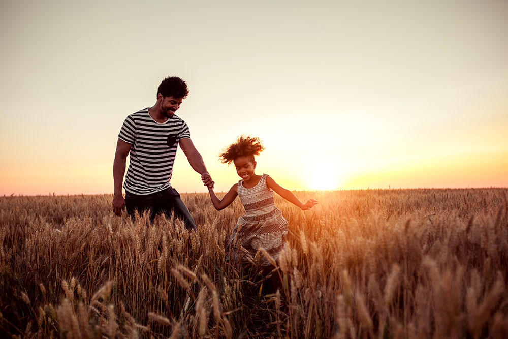 Father and daughter are running and jumping through a field of wheat during the golden hour of sunset