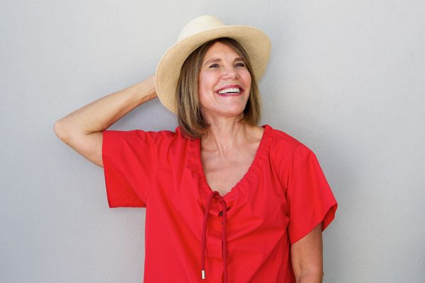 older woman smiling in a bright read shirt with her hand on her hat