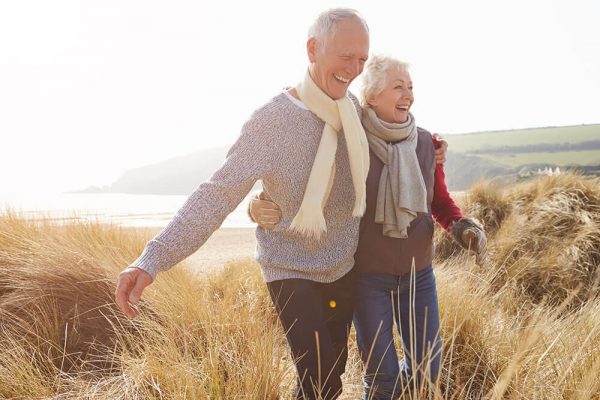Older couple walking through grass, with their arms around each other, with a beach scene in the background